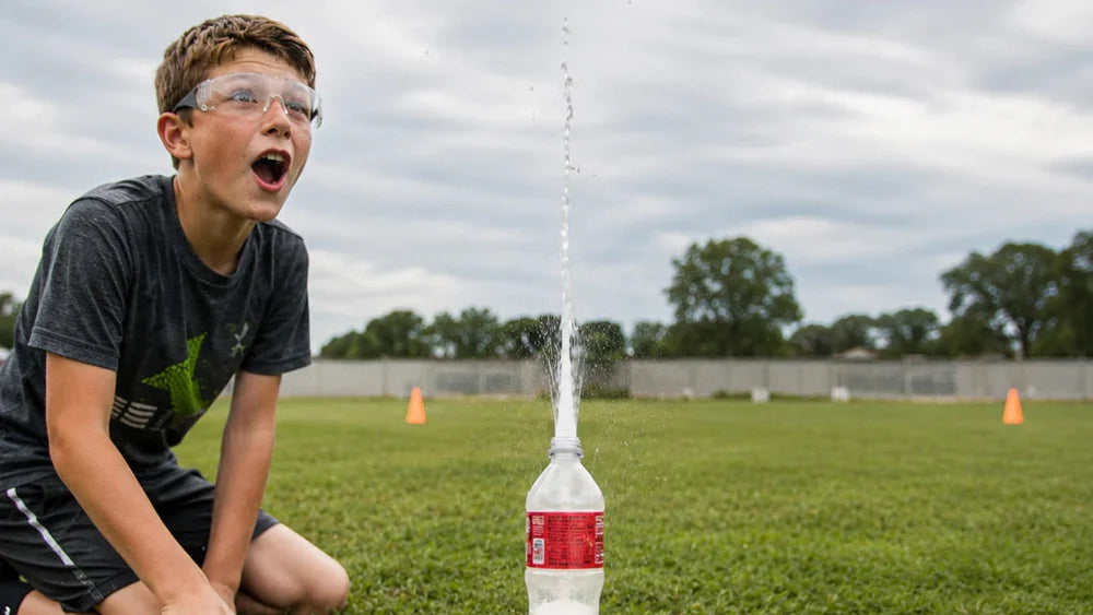 A boy wearing safety goggles crouches on a grassy field, looking up in surprise at a water fountain shooting upwards from a clear plastic bottle with a red label.
